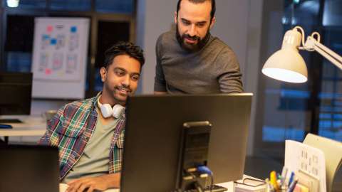 Dos compañeros de trabajo en un espacio de trabajo moderno y abierto revisan juntos la información que se muestra en un monitor de un equipo de desktop. Una persona está sentada y sonríe mientras escribe en un teclado y la otra se encuentra de pie y señala algo en la pantalla.