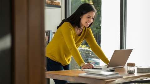 Una persona de pie en una oficina en casa se inclina hacia adelante y apoya ambas manos en el escritorio mientras sonríe a la pantalla de una laptop abierta